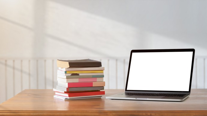 Auf dem Bild sehen wir einen Tisch auf dem sich links ein Stapel Bücher und daneben ein aufgeklappter Laptop mit weißem Bildschirm befindet Auf dem Bild sehen wir einen Tisch auf dem sich links ein Stapel Bücher und daneben ein aufgeklappter Laptop mit weißem Bildschirm befindet