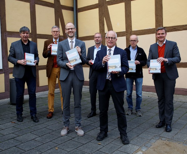 Sieben Herrn präsentieren vor einem Fachwerkhaus das Buch
